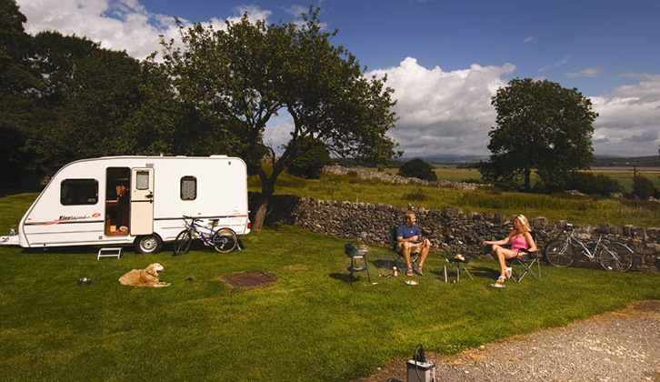 A couple sitting on chairs with their dog and caravan nearby on a bright day