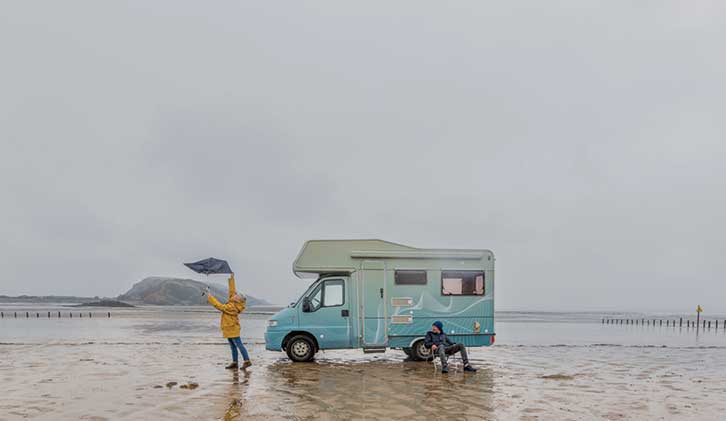 Person on beach holding umbrella up on grey day by motorhome