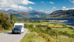 A car towing a caravan along a road with Ullapool and Loch Broom in Scotland visible in the background