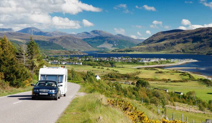 A car towing a caravan along a road with Ullapool and Loch Broom in Scotland visible in the background