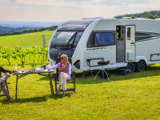A couple eating and drinking at a camping table by a Conqueror caravan