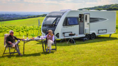 A couple eating and drinking at a camping table by a Conqueror caravan