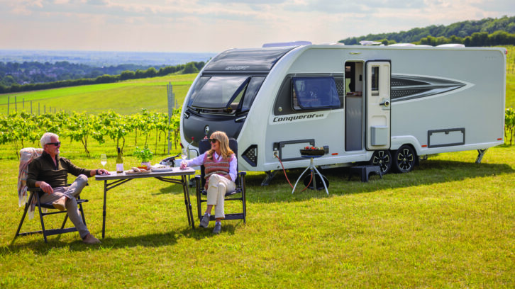 A couple sitting eating and drinking at a camping table by a Conqueror caravan