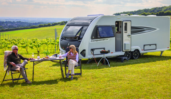 A couple eating and drinking at a camping table by a Conqueror caravan