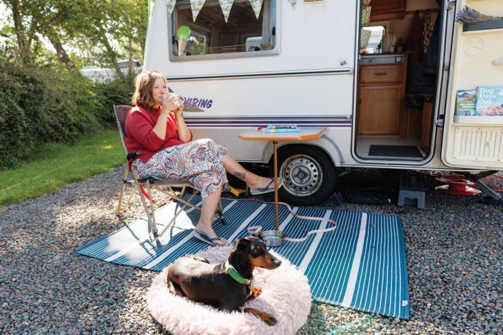 Lottie sitting back in a camping chair drinking a cup of tea while Arty lies down on a rug